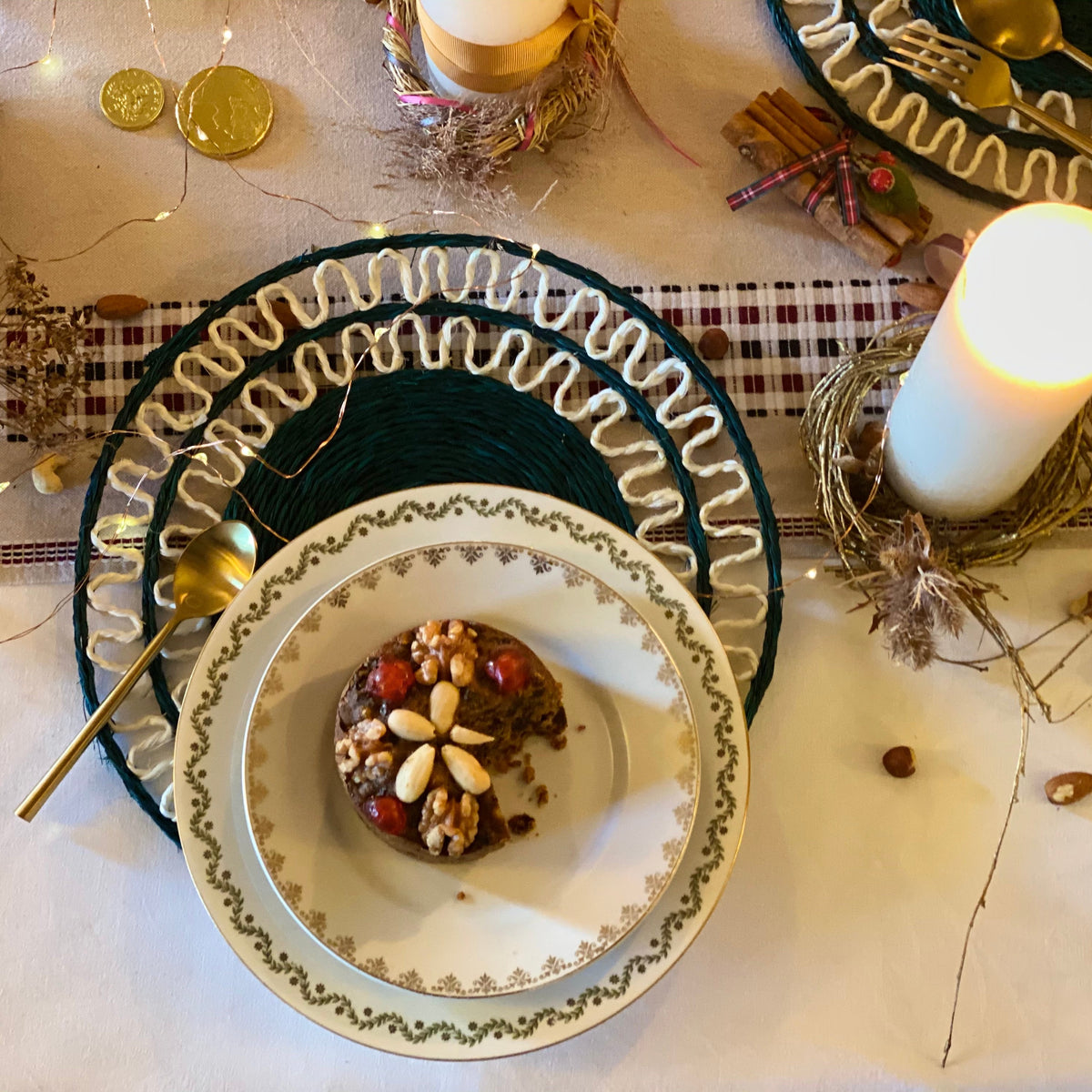 A styled holiday table setting featuring a white ceramic plate with a gold floral border nested inside a dark green ruffled charger plate. A small decorated nut and dried fruit dessert sits on the plate, accompanied by a gold spoon, with a lit white pillar candle and rustic holiday decorations in the background.